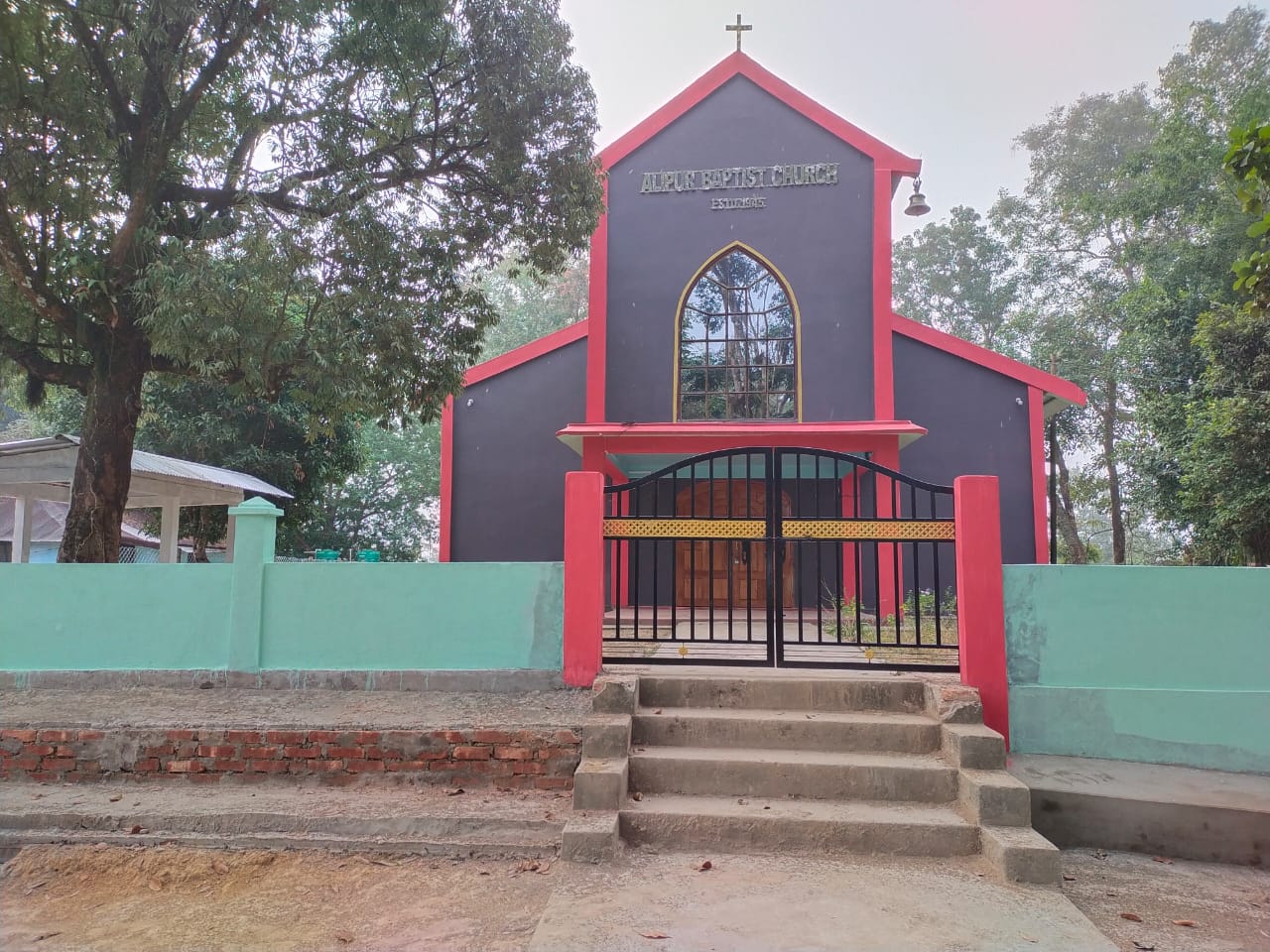 The Alipur Baptist Church building in Banskandi, framed by trees and its green entrance wall